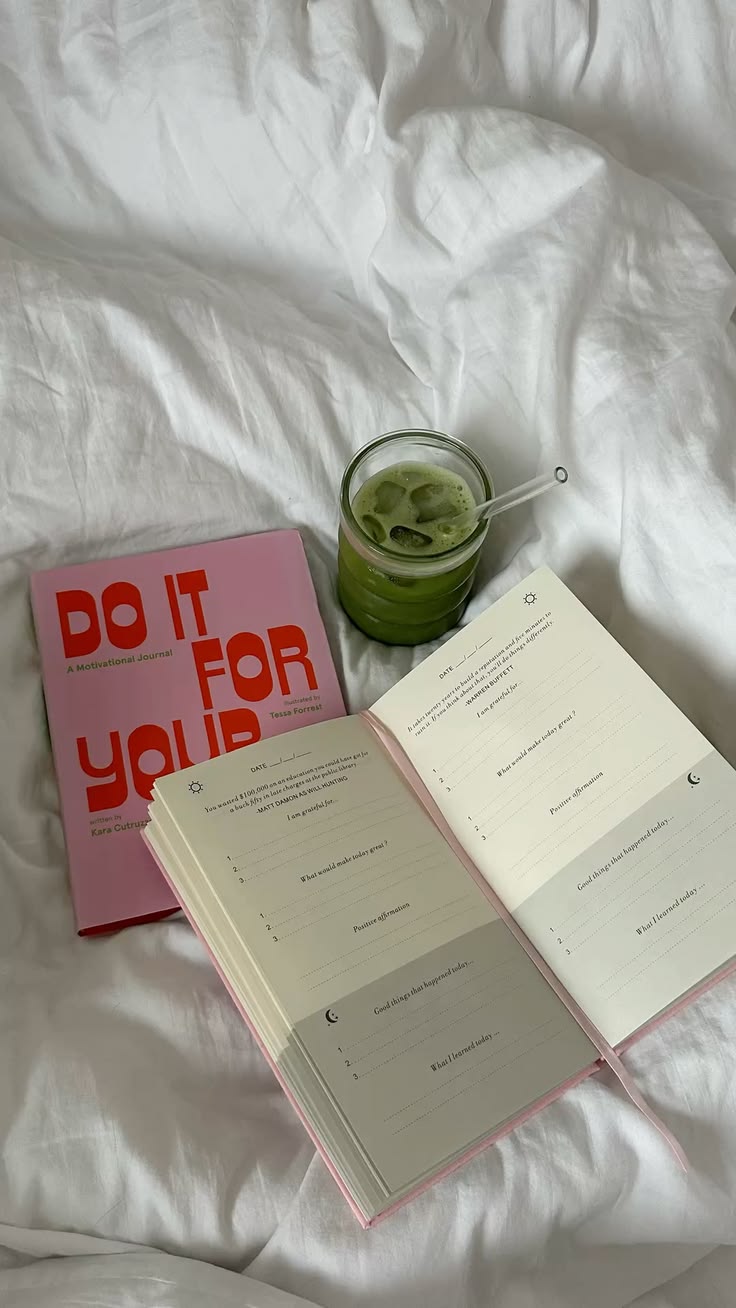 Pink self-help book, open journal, and a glass of green iced drink on a white bedsheet.