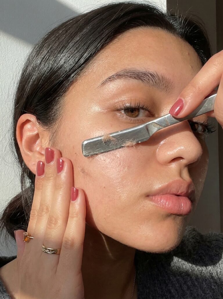 A woman performs at home dermaplaning with a facial razor, exfoliating her skin for smooth, glowing results using skincare tools.