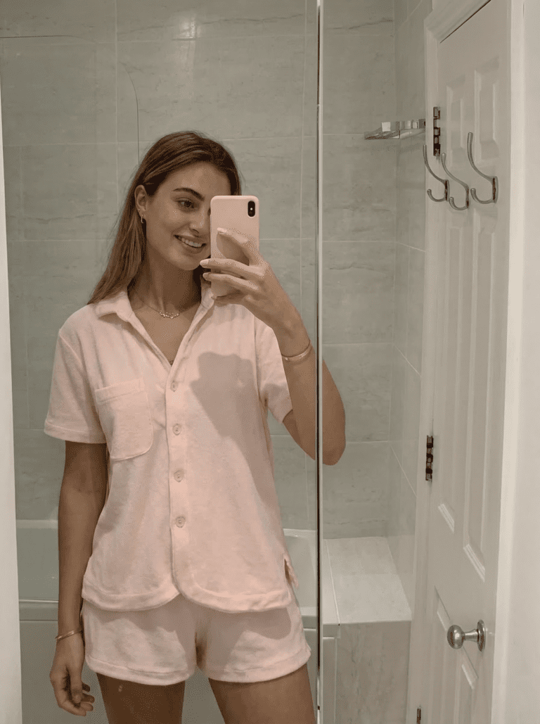 Woman wearing cute pink pajamas snapping a mirror selfie in a modern beige tile bathroom for fashion and loungewear trends.