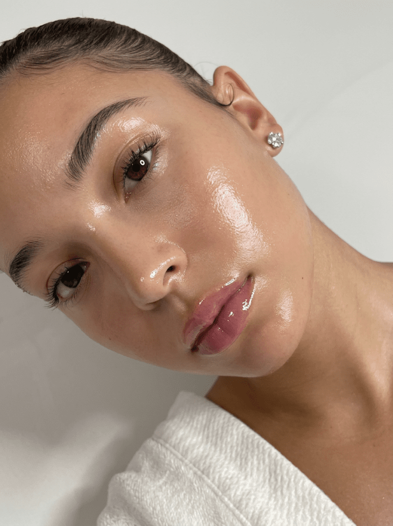 Close-up of woman with glass skin, radiant dewy complexion, glossy lips, diamond earring, white background—K-beauty trend.