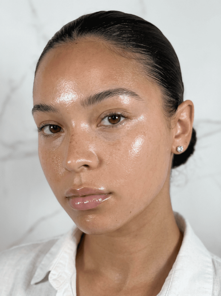 Woman with radiant glass skin, dewy complexion, slicked-back hair, stud earrings, and a white button-up shirt for skincare routine.