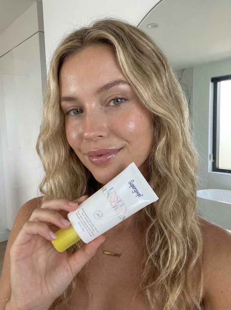 Smiling woman with wavy blonde hair in bright bathroom holding sunscreen for oily skin, skincare routine for glowing complexion.