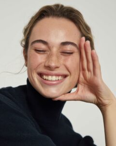 Happy woman relaxing, smiling with eyes closed, hand on face, wearing cozy dark turtleneck sweater, peaceful and content.
