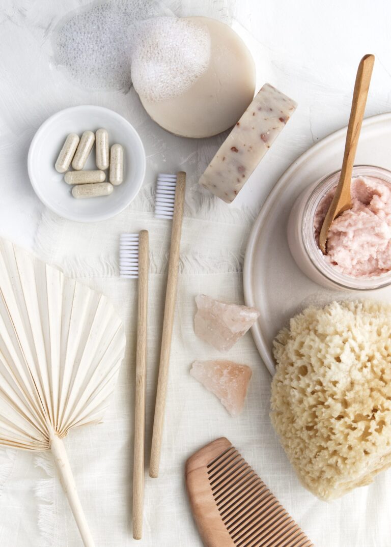 Natural spa essentials flat lay with organic soap bars, exfoliating scrub jar, bamboo toothbrushes, eco sponge, wooden comb, supplements.