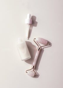 Anti-aging facial serum bottle with dropper and rose quartz face roller on pastel pink background, skincare beauty flatlay.