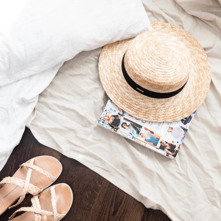 Summer essentials: straw hat, beach sandals, and fashion magazines on white sheet near dark hardwood flooring, vacation flat lay.