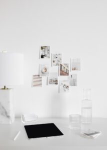 Modern home office workspace with minimalist white desk, tablet, glass of water, stylish lamp, and aesthetic photo wall decor.