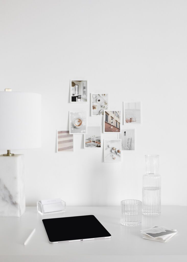 Modern home office workspace with minimalist white desk, tablet, glass of water, stylish lamp, and aesthetic photo wall decor.