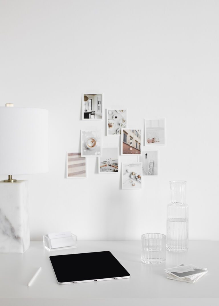 Modern home office workspace with minimalist white desk, tablet, glass of water, stylish lamp, and aesthetic photo wall decor.