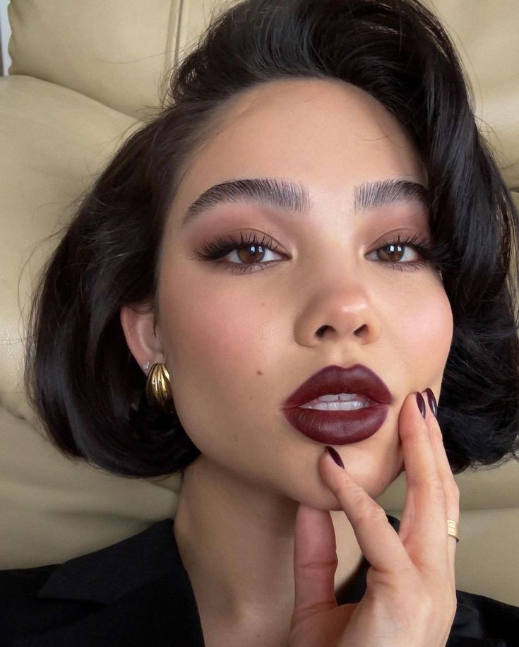 Chic woman selfie with short dark hair, burgundy lipstick, gold hoop earrings, stylish manicure, beauty influencer look.