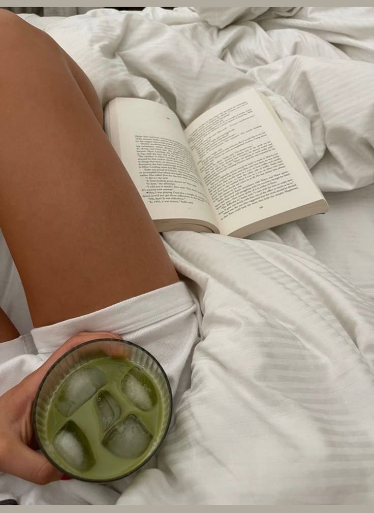 Woman reading a book on a cozy white bed, holding an iced matcha latte, relaxing bedroom decor, peaceful morning routine.