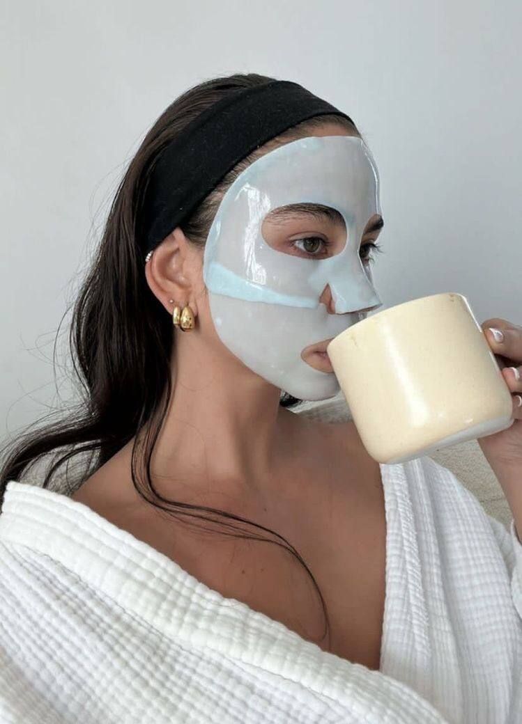 woman in white robe sipping coffee with face mask on