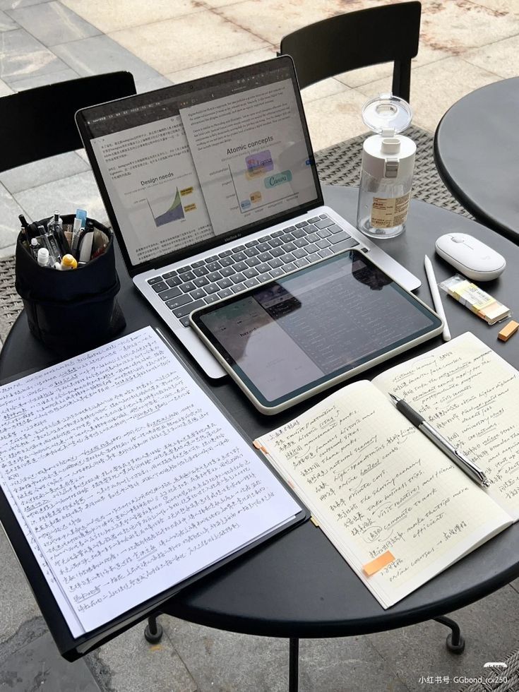 A table with a laptop, tablet, notebook, handwritten notes, pens, and a bottle of drink arranged for studying.